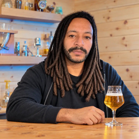 Josh O, Craft a Brew’s Fulfillment Manager, seated at a wooden table with a stemmed glass of beer.