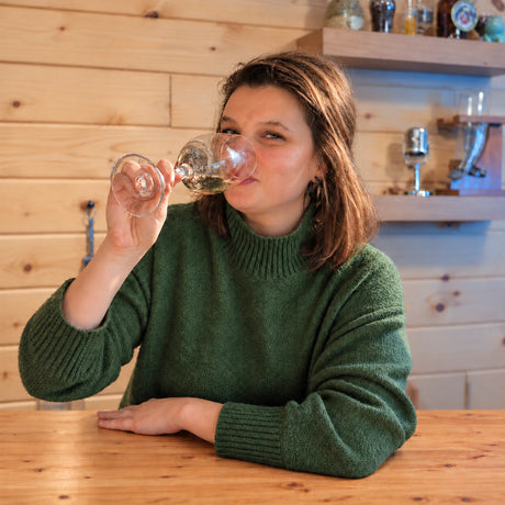 Kaley K, Craft a Brew’s Customer Experience & Marketing Manager, sipping from a stemmed glass at a wooden table.