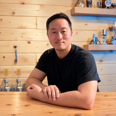 David Kim, Craft a Brew’s Ingredient Manager, seated at a wooden table with glassware displayed behind him.