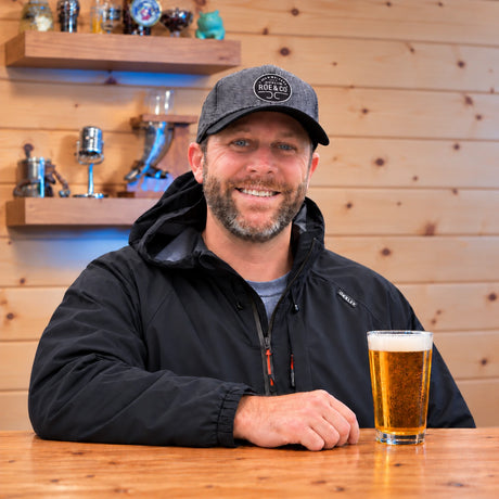 Kyle Westfall, founder of Craft a Brew, smiling at a wooden table with a pint of beer.